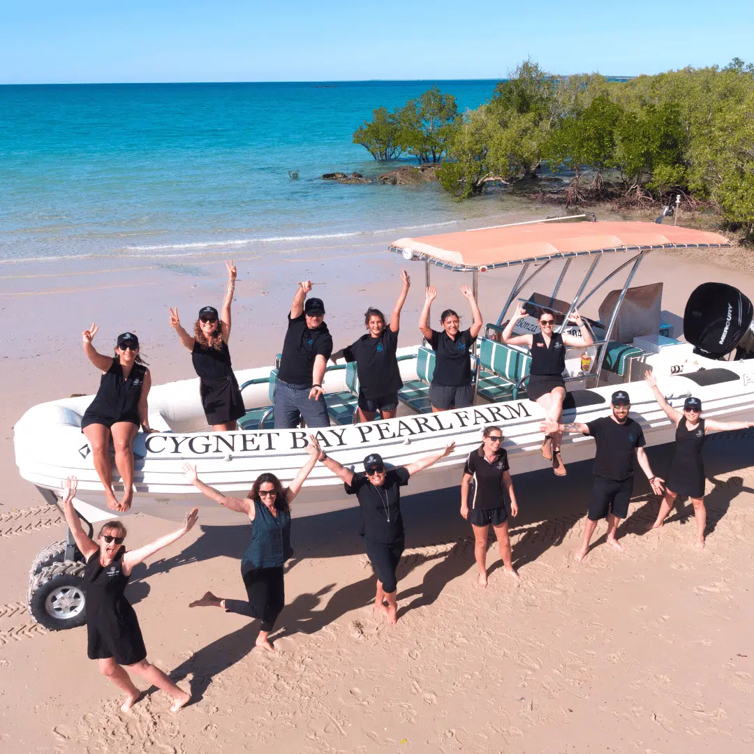 Our Cygnet Bay Pearl Farm team surround a boat used for tours, all posing and smiling while stand on the beach. In the background is the iconic blue waters of the Kimberley Coastline.
